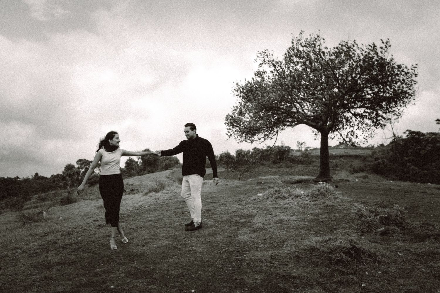 A couple walks together on a hillside with a lone tree in the distance.