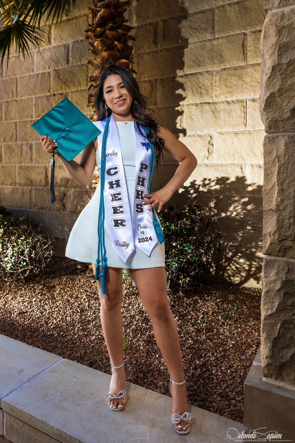Graduate in teal gown and white stole poses against stone wall.