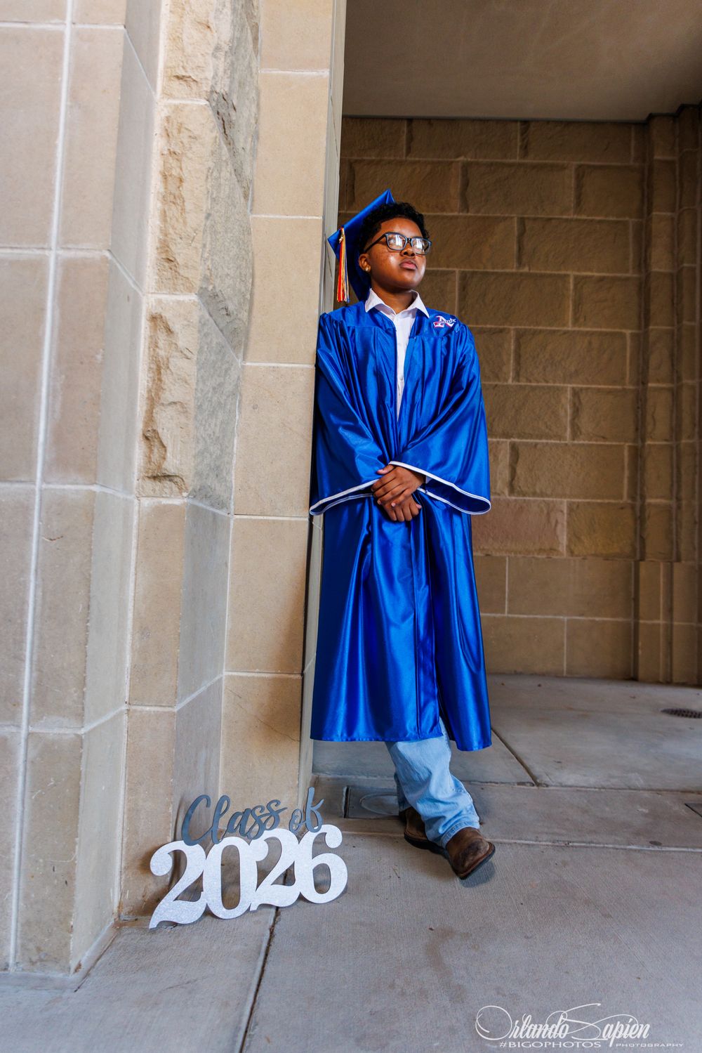 A student in a blue graduation gown and cap poses by a stone wall with a Class of 2026 sign.