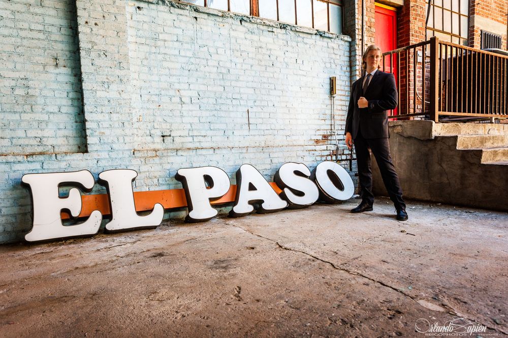 Man in suit posing beside large white El Paso letter signs against a brick wall backdrop.