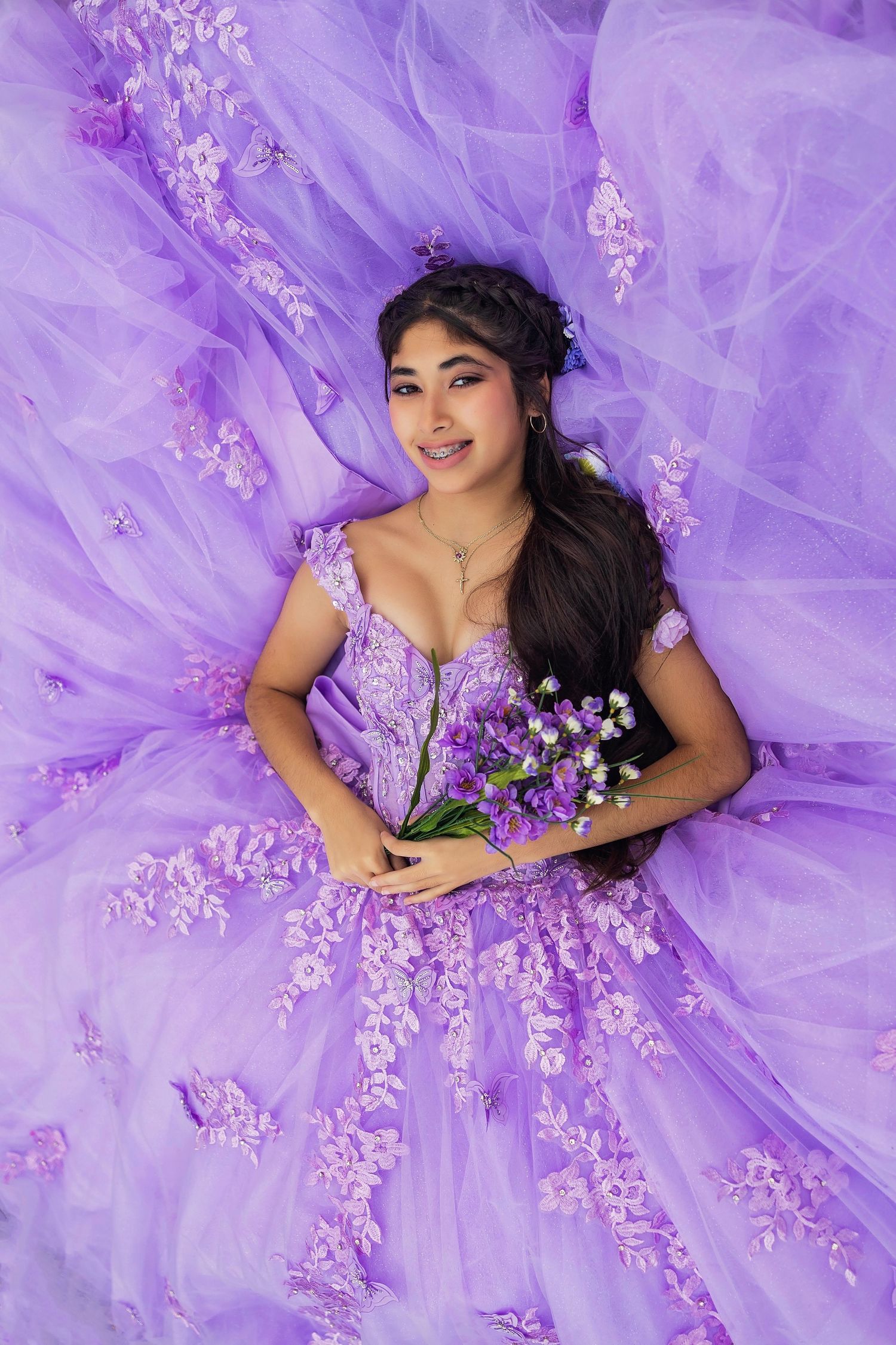 Smiling young woman in a lavender quinceañera ball gown with floral appliqués lying on tulle, holding a bouquet of purple flowers.