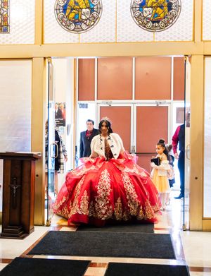 Quinceañera in a red and gold ball gown with a tiara enters a church doorway as family members watch near stained glass windows.