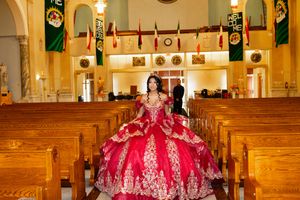 A quinceañera in a sparkling red and gold ball gown stands in the aisle of a church lined with wooden pews and banners.