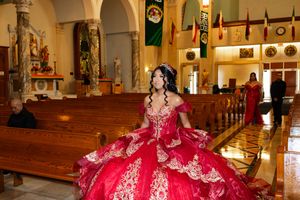 Quinceañera in a red and gold embellished ball gown with a tiara posing in a church aisle beside wooden pews and Mexican flags.