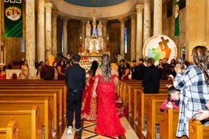 Quinceañera in a long red gown walks down a church aisle between wooden pews toward the ornate altar.