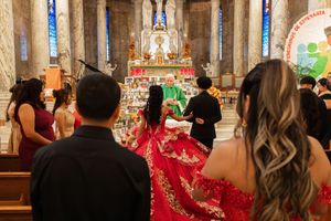 Quinceañera in a red embroidered gown stands before a church altar as guests and a priest in green vestments gather for her Mass.
