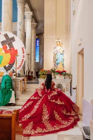 A quinceañera in a red and gold embroidered gown kneels before a Virgin Mary statue at a church altar during her ceremony.