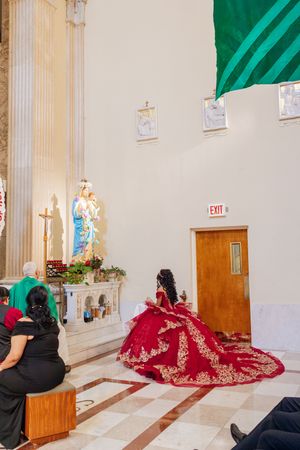 Young woman in an elaborate red quinceañera gown kneels before a Virgin Mary statue inside a church while family members watch.