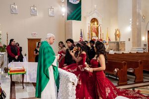 A priest in green vestments stands at a church altar blessing a quinceañera in a red gown, with family gathered beside her.