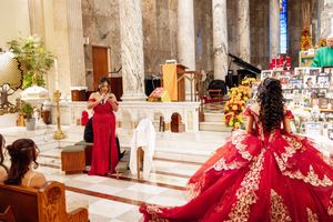 Quinceañera in a voluminous red embroidered ball gown kneels in a church sanctuary near flowers and a priest.