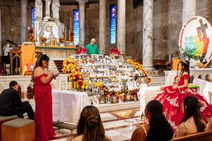 A quinceañera in a red ball gown kneels near a flower-covered memorial altar with photos inside a church as relatives look on.