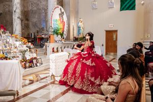 A quinceañera in a sparkling red ball gown poses beside an altar with flowers and photos during a church celebration as guests look on.