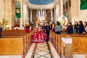 Quinceañera in a red gown walks down the aisle of a church with an escort during a Mass celebration.