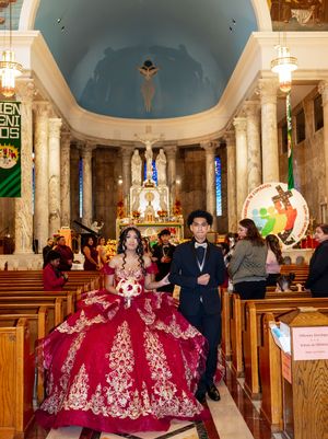 A young woman in a red embroidered quinceañera ball gown poses with a young man in a black tuxedo inside a church.