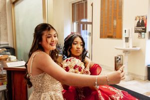 Two young women in formal dresses take a selfie together, one in a red gown with a tiara holding a bouquet of red roses.