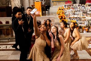 A group of quinceañera court members in gold dresses and black suits pose for a selfie inside a church sanctuary.
