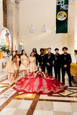 A quinceañera in an ornate red and gold gown poses on a church floor with her court of damas and chambelanes in formal attire.
