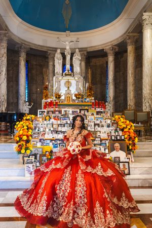 Quinceañera in a red gown with gold embroidery holding a bouquet while posing before a church altar with photos and marigold flowers.