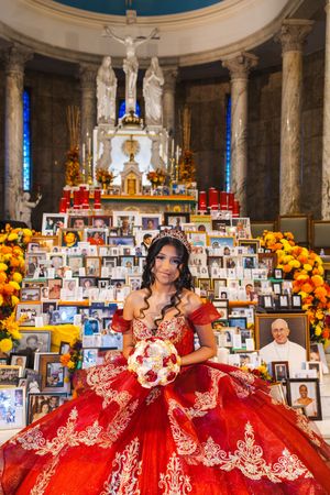 Smiling quinceañera in a red embroidered gown holds a bouquet before a church altar surrounded by framed photos and flowers.