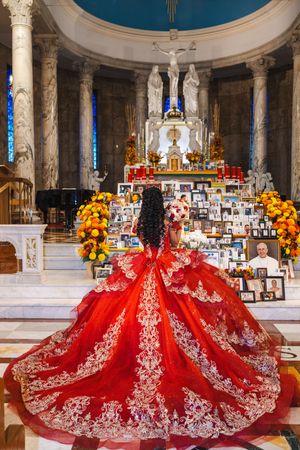 Quinceañera in a red ball gown with gold embroidery faces a church altar decorated with marigolds and framed photographs.