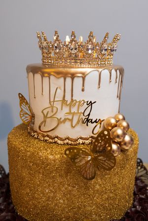 Two-tier birthday cake with gold drip icing, a jeweled crown topper, gold butterflies and glitter, and a Happy Birthday sign.