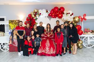 Family gathered around a girl in a red quinceañera gown, posing in front of red, white and gold balloon decorations at a celebration.