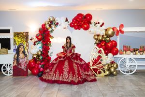 Quinceañera in a red and gold embroidered ball gown posing before a balloon arch with a portrait display and decorated carts.