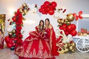 Two women pose in red and gold gowns, one in a quinceañera ball gown, beside red and gold balloon arches, flowers, and a decorated cart.