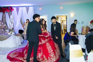 Quinceañera in a sparkling red ball gown stands with a man in a black suit while guests mingle in a decorated banquet hall.
