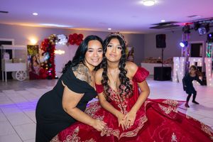 Two women pose together at a quinceañera party, one in an elegant red beaded gown and tiara, with festive balloons and lights behind.