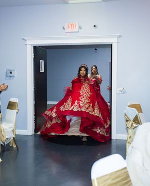 Young woman in a voluminous red quinceañera ball gown with gold embroidery and a tiara entering a banquet hall, with a guest behind her.