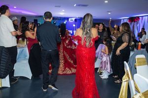 Guests gather on a dance floor at an evening celebration, with a woman in a long red sparkly gown standing at the center.
