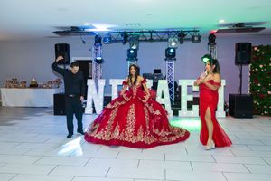Quinceañera in a red embellished ball gown poses on a dance floor with a woman in a red dress and a DJ beside large illuminated letters.
