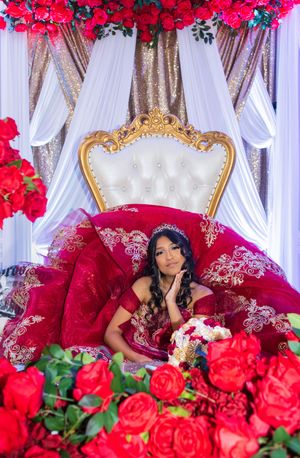 Quinceañera in a red embroidered ball gown and tiara sits on a gold throne surrounded by red roses and white draping.