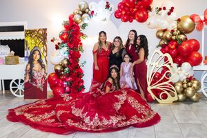 Quinceañera in a red ball gown poses with family beside red and gold balloon decor, a butterfly wing prop, and a framed portrait.