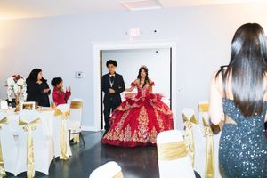 Quinceañera in a sparkling red and gold ball gown with a tiara enters a decorated banquet hall as guests in white chairs watch.