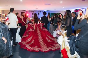 A quinceañera in a voluminous red and gold embroidered ball gown stands among cheering guests holding up phones at a party.