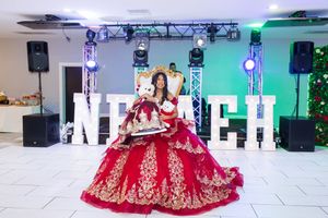 Girl in a red and gold embellished quinceañera ball gown holding a teddy bear, seated before large illuminated letters at a party venue.