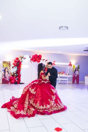 A quinceañera in a red and gold embroidered ball gown dances with a man in black near a red balloon arch and dessert table.