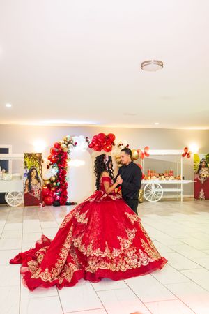 A quinceañera in a red and gold embroidered ball gown dances with a man in black near a balloon arch and dessert cart.