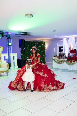 Quinceañera in an ornate red and gold ball gown dances with a young girl in white at a decorated banquet hall.