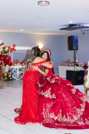 Two women in elegant red dresses embrace at a quinceañera celebration, with a dessert table and balloon decor in the background.