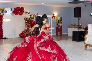 Quinceañera in a red embroidered ball gown dances with a loved one in a festive hall decorated with red and gold balloons.
