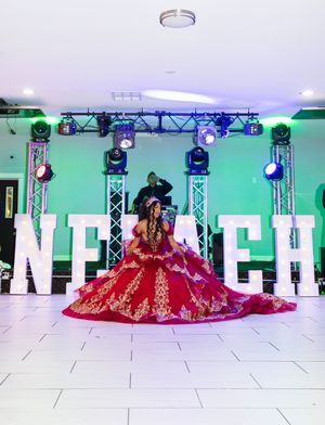 Quinceañera in a red and gold embroidered ball gown posing on a white tile dance floor before light-up letters and a DJ booth.