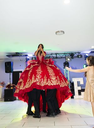 Quinceañera in a red gown with gold embroidery lifted high on a hidden platform by dancers while a guest reaches up on the dance floor.
