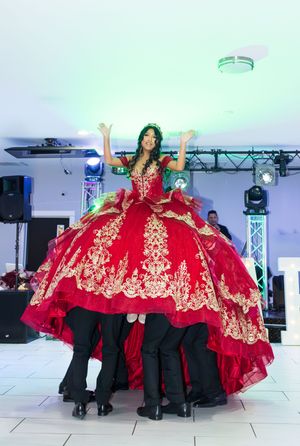 Quinceañera in a red and gold embroidered ball gown raises her arms while lifted high by escorts in black suits at a lively party.
