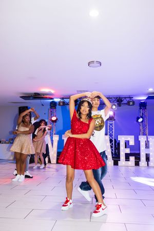 Girl in a sparkly red dress and tiara dances with a boy on a lit dance floor at a quinceañera party while friends dance behind them.