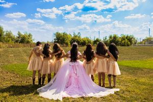 Quinceañera in a flowing pink ball gown stands with her court in peach dresses, all facing a grassy field under a blue sky.