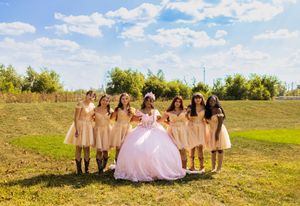 Quinceañera in a pink ball gown poses with seven damas in champagne dresses and boots on a grassy hillside under a blue sky.