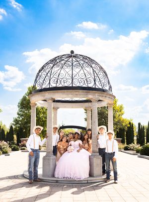 Quinceañera in a pink ball gown poses with her court inside an ornate stone gazebo, flanked by men in white shirts and straw hats.